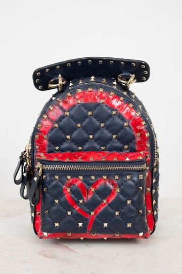 A small quilted backpack with navy blue and red details, gold-tone studs, and a red heart design on the front, photographed against a plain light background.