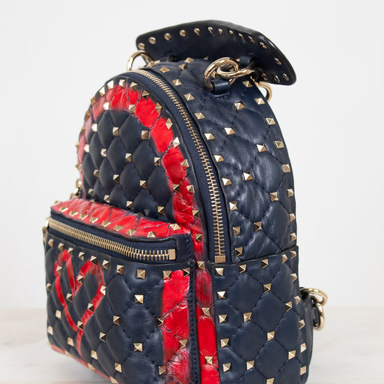 A small quilted backpack in navy blue and red, decorated with gold pyramid studs and featuring a red heart motif on the front pocket, set against a plain light background.