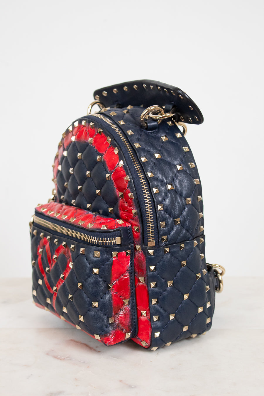 A small quilted backpack in navy blue and red, decorated with gold pyramid studs and featuring a red heart motif on the front pocket, set against a plain light background.