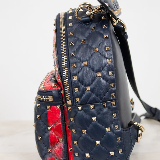 A navy blue quilted leather backpack with gold pyramid studs, featuring a red and black floral patterned fabric lining, set against a light gray background.