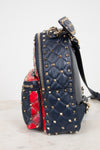 A navy blue quilted leather backpack with gold pyramid studs, featuring a red and black floral patterned fabric lining, set against a light gray background.