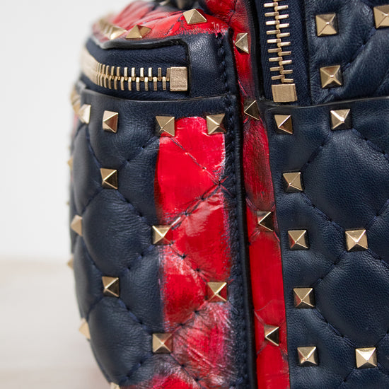 Close-up of a quilted leather bag in navy blue with gold pyramid studs and a bold red quilted accent panel, showing a gold zipper.