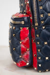 Close-up of a quilted leather bag in navy blue with gold pyramid studs and a bold red quilted accent panel, showing a gold zipper.