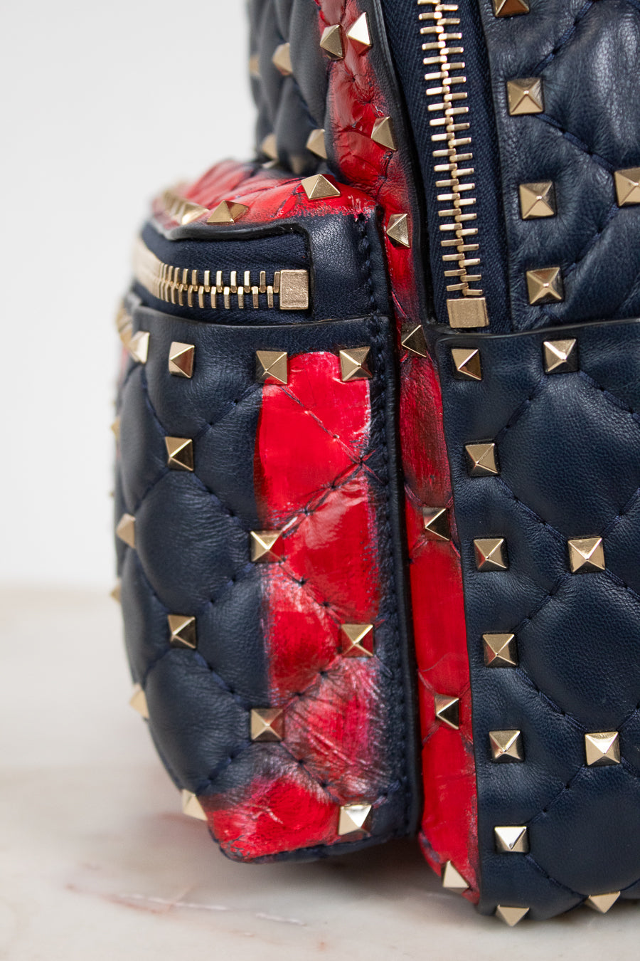 Close-up of a quilted leather bag in navy blue with gold pyramid studs and a bold red quilted accent panel, showing a gold zipper.
