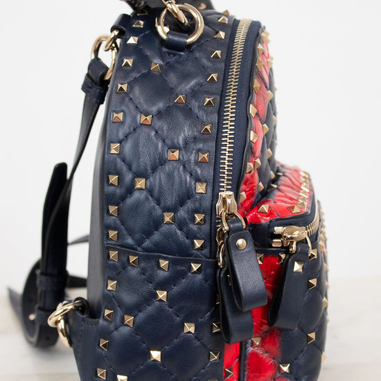 A navy blue quilted leather backpack with gold pyramid studs, featuring a red and navy patterned print side panel and gold-tone zippers.