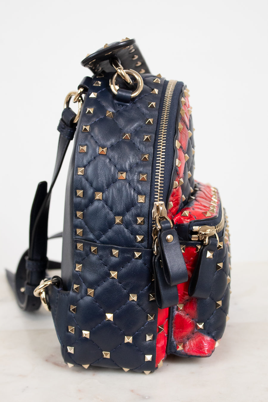 A navy blue quilted leather backpack with gold pyramid studs, featuring a red and navy patterned print side panel and gold-tone zippers.