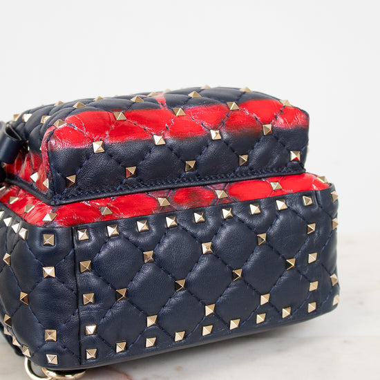 A navy blue quilted leather handbag with gold pyramid studs and a vibrant red leather top flap details, shown on a light marble surface against a white background.