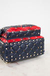 A navy blue quilted leather handbag with gold pyramid studs and a vibrant red leather top flap details, shown on a light marble surface against a white background.
