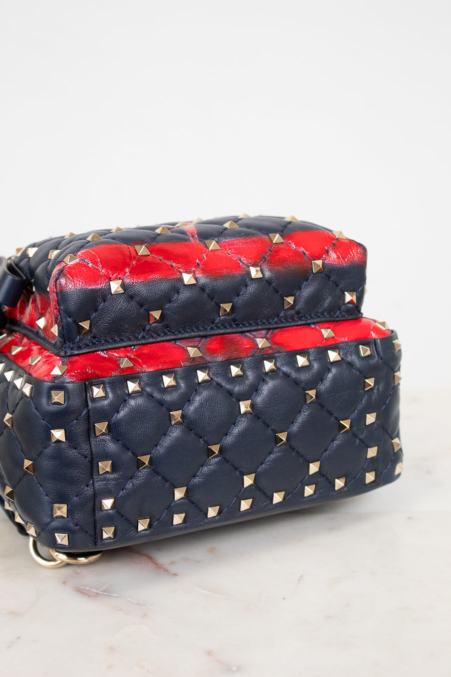 A navy blue quilted leather handbag with gold pyramid studs and a vibrant red leather top flap details, shown on a light marble surface against a white background.