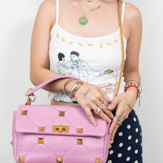 A woman wearing a white graphic camisole and navy polka-dot pants holds a pink quilted shoulder bag with gold-tone studs and chain strap against a plain light background.