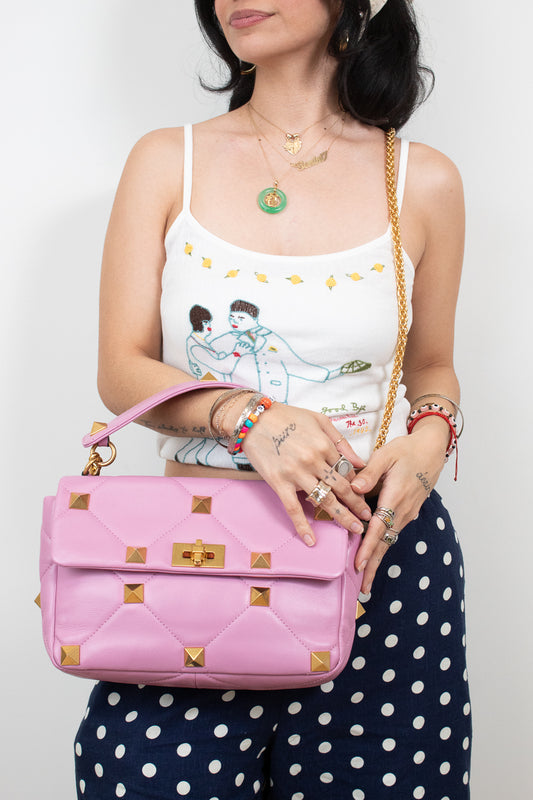 A woman wearing a white graphic camisole and navy polka-dot pants holds a pink quilted shoulder bag with gold-tone studs and chain strap against a plain light background.