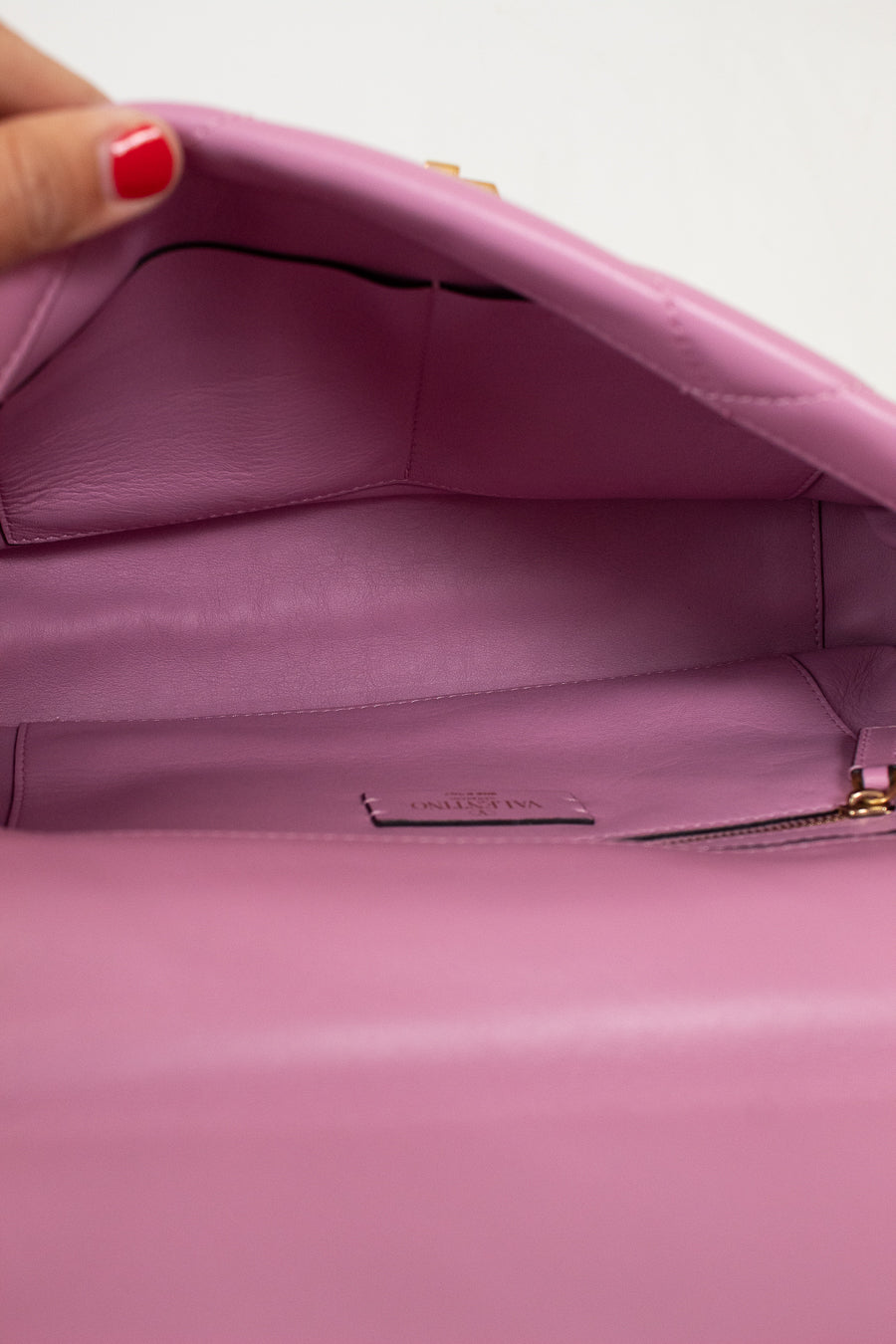 Close-up of the interior of a pink leather handbag, showing a zippered compartment and a small interior label, with a hand holding the open flap at the top left.