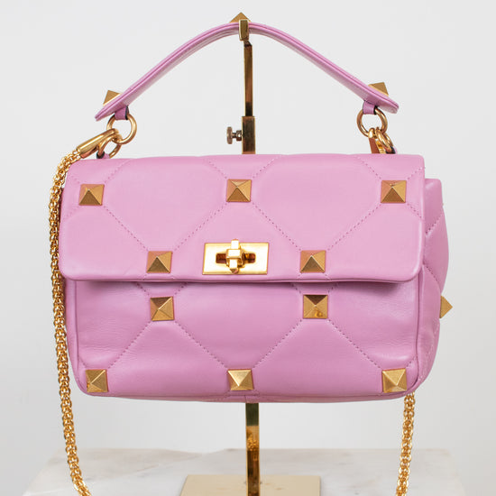 A pink quilted leather handbag with gold pyramid studs and a gold chain strap, displayed on a gold stand against a white background.