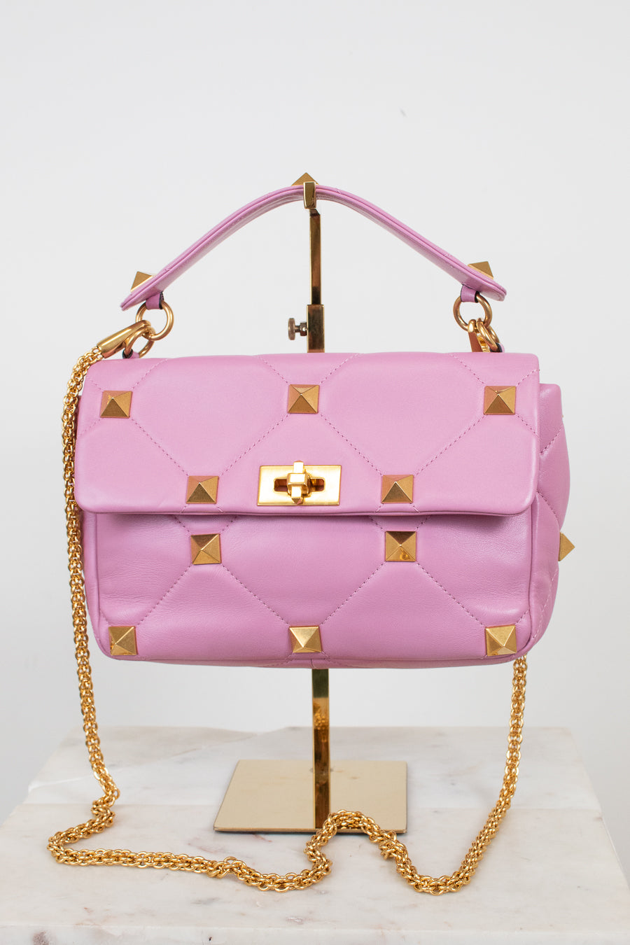 A pink quilted leather handbag with gold pyramid studs and a gold chain strap, displayed on a gold stand against a white background.