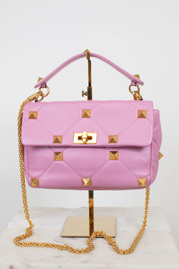 A pink quilted leather handbag with gold pyramid studs and a gold chain strap, displayed on a gold stand against a white background.