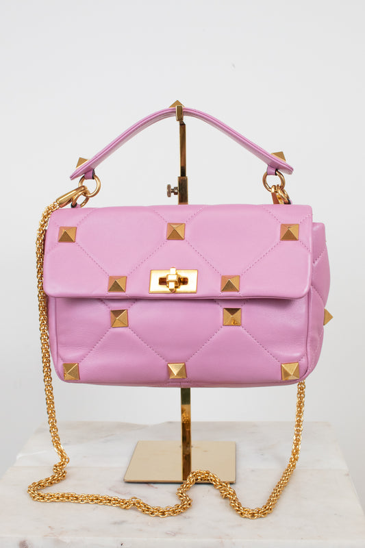 A pink quilted leather handbag with gold pyramid studs and a gold chain strap, displayed on a gold stand against a white background.