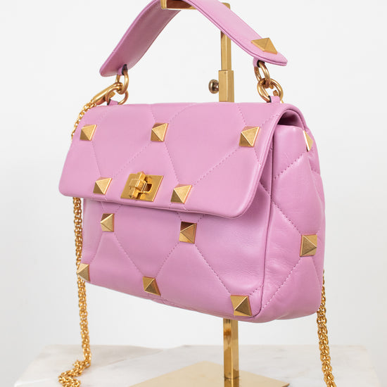 A pink quilted leather handbag with gold pyramid studs and a gold chain strap, displayed on a gold stand against a white background.