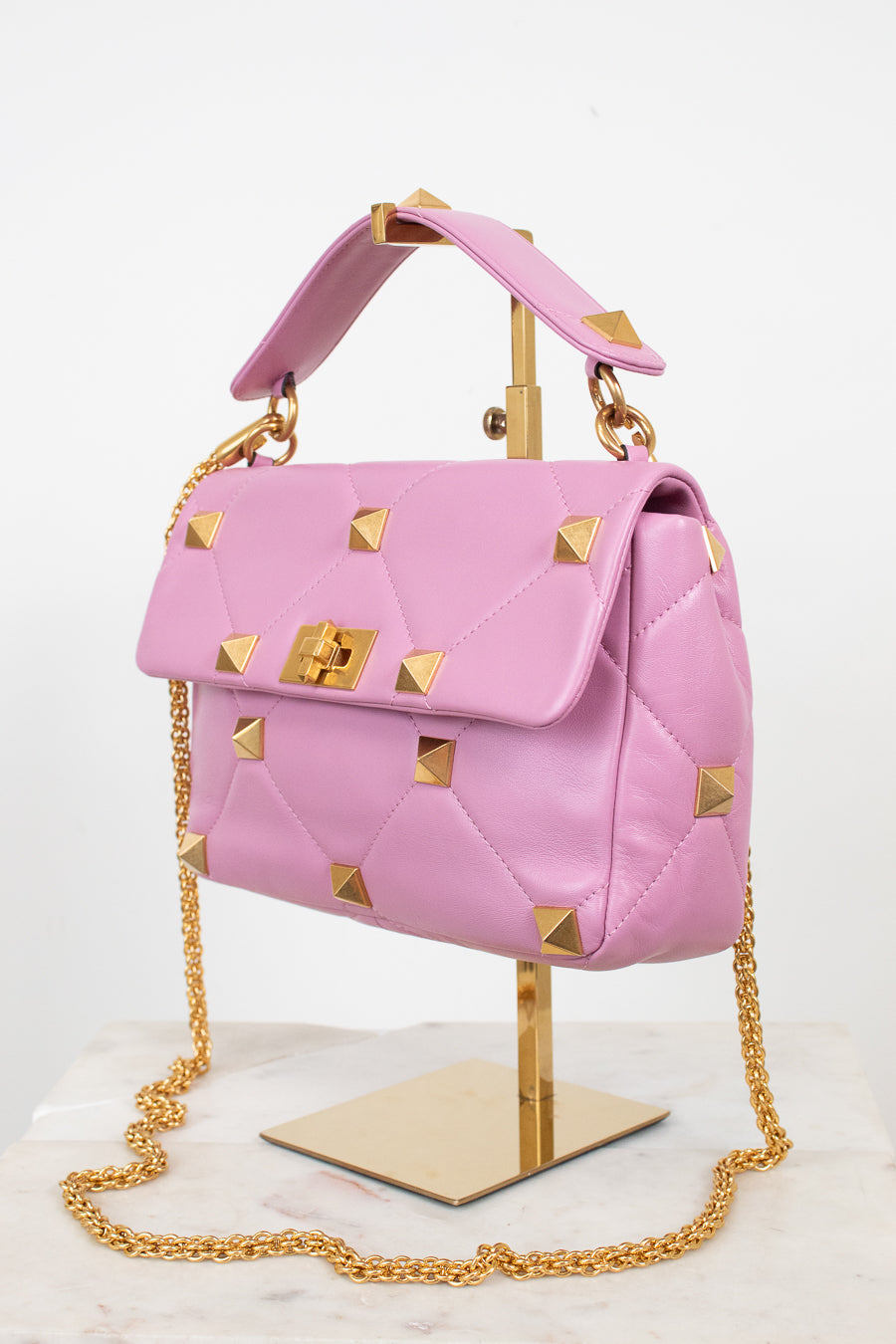 A pink quilted leather handbag with gold pyramid studs and a gold chain strap, displayed on a gold stand against a white background.