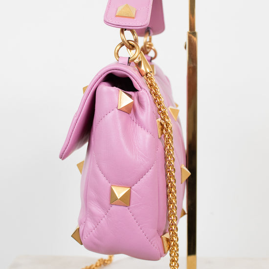 A pink quilted leather handbag with gold pyramid studs and a gold chain strap, displayed on a gold stand against a white background.