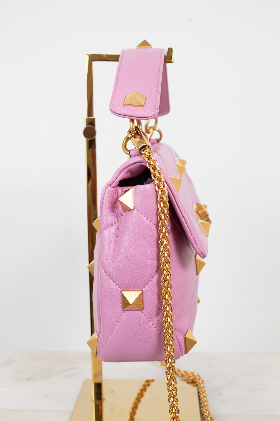 A pink quilted leather handbag with pyramid studs and a gold chain strap, displayed on a gold stand against a white background.