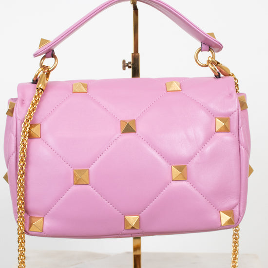 Pink quilted leather handbag with gold pyramid studs and a gold chain strap, displayed on a gold stand against a white background.