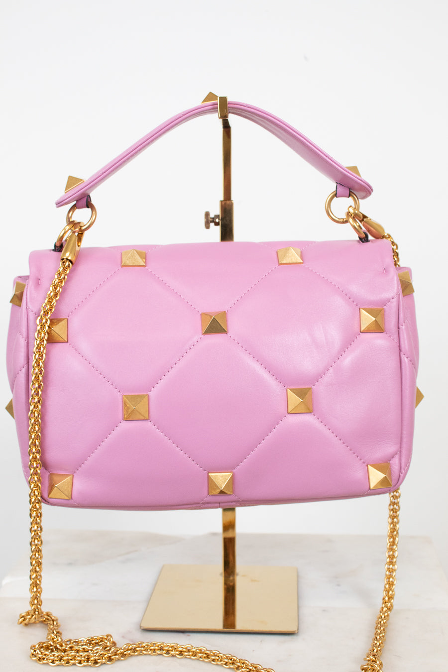 Pink quilted leather handbag with gold pyramid studs and a gold chain strap, displayed on a gold stand against a white background.