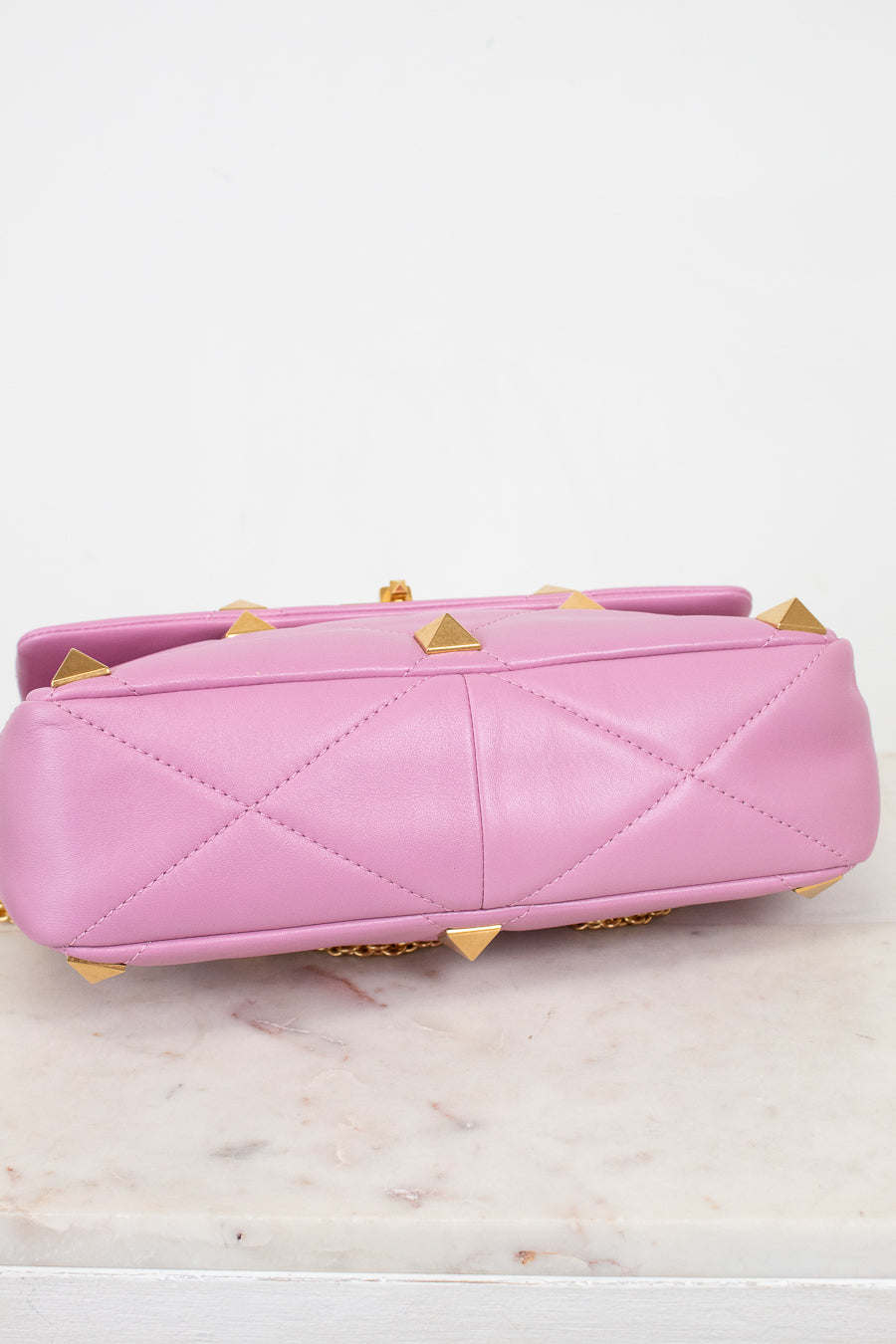 A pink quilted leather handbag with gold pyramid studs, shown from the bottom and laying on a light marble surface against a white background.