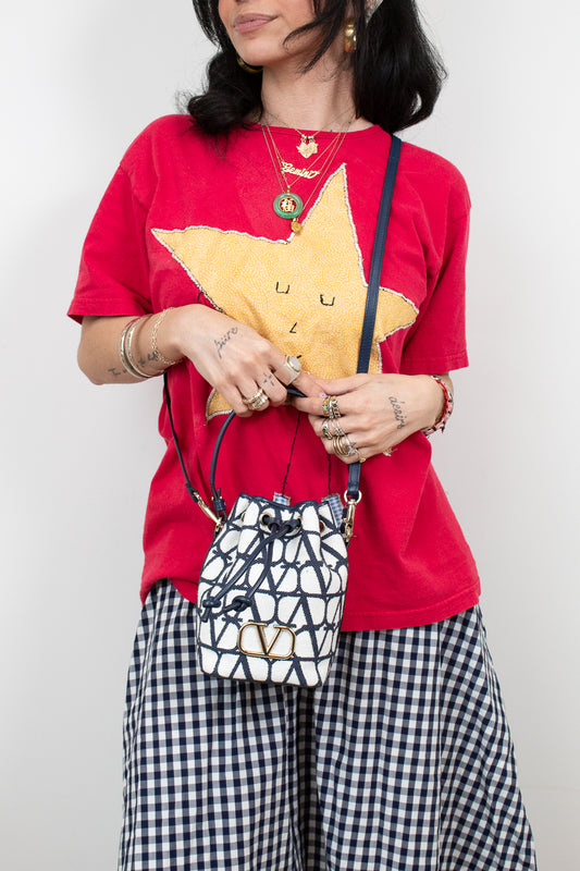Person wearing a red shirt with a yellow star design, holding a patterned handbag against a plain background.