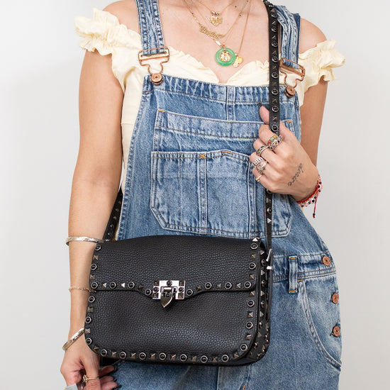 Person wearing denim overalls holding a black studded handbag against a plain background