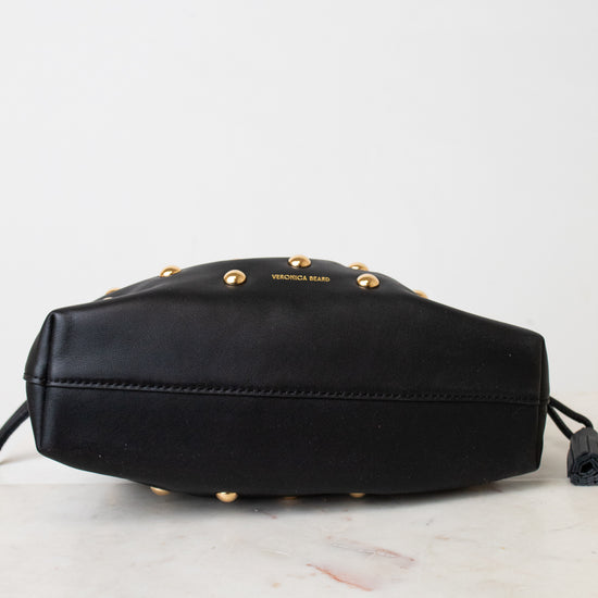 A black leather bag with gold-tone studs and the brand name 'VERONICA BEARD' printed on the front, resting on a white marble surface against a white background.