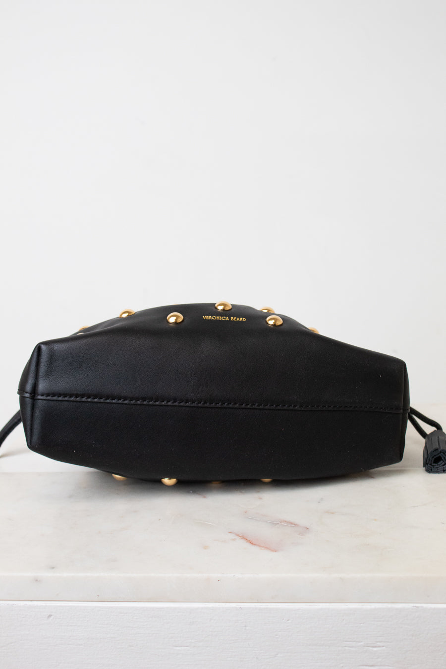 A black leather bag with gold-tone studs and the brand name 'VERONICA BEARD' printed on the front, resting on a white marble surface against a white background.