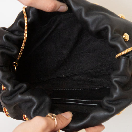 A close-up of a black leather bag being held open by two hands wearing rings, showing the gold-tone chain strap and gold studs.