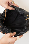 A close-up of a black leather bag being held open by two hands wearing rings, showing the gold-tone chain strap and gold studs.