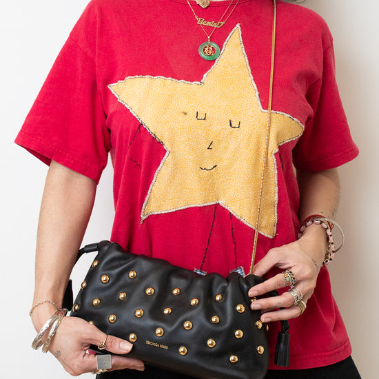 A woman wearing a red graphic t-shirt with a large yellow star design, black shorts, and holding a black studded leather shoulder bag with a gold chain strap.