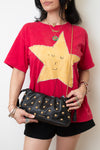A woman wearing a red graphic t-shirt with a large yellow star design, black shorts, and holding a black studded leather shoulder bag with a gold chain strap.