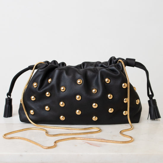 Black leather drawstring clutch bag with gold-tone studs, black tassels, and a gold chain shoulder strap, displayed on a white surface against a white background.