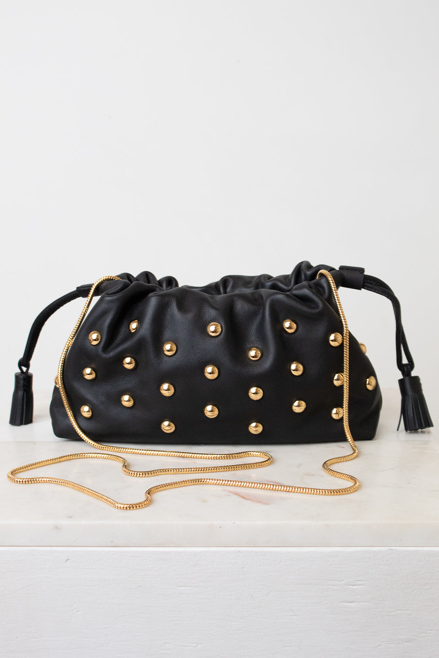 Black leather drawstring clutch bag with gold-tone studs, black tassels, and a gold chain shoulder strap, displayed on a white surface against a white background.