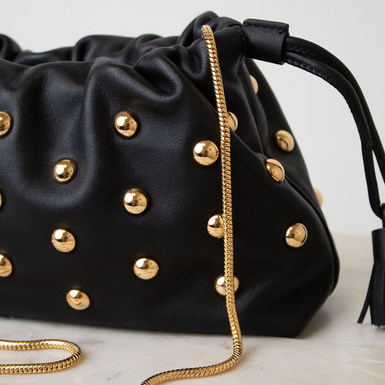 A black leather drawstring handbag adorned with gold-tone studs and a gold chain strap, resting on a light marble surface against a neutral light gray background.