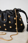 A black leather drawstring handbag adorned with gold-tone studs and a gold chain strap, resting on a light marble surface against a neutral light gray background.