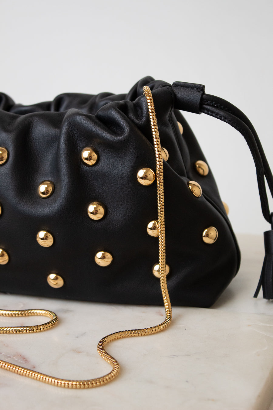 A black leather drawstring handbag adorned with gold-tone studs and a gold chain strap, resting on a light marble surface against a neutral light gray background.