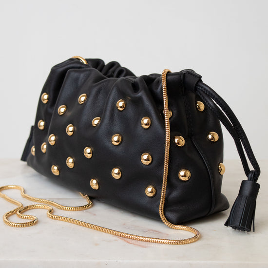 Black leather clutch bag with gold-tone studs, a gold chain strap, and a black tassel detail, displayed on a white surface against a light gray background.