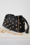 Black leather clutch bag with gold-tone studs, a gold chain strap, and a black tassel detail, displayed on a white surface against a light gray background.