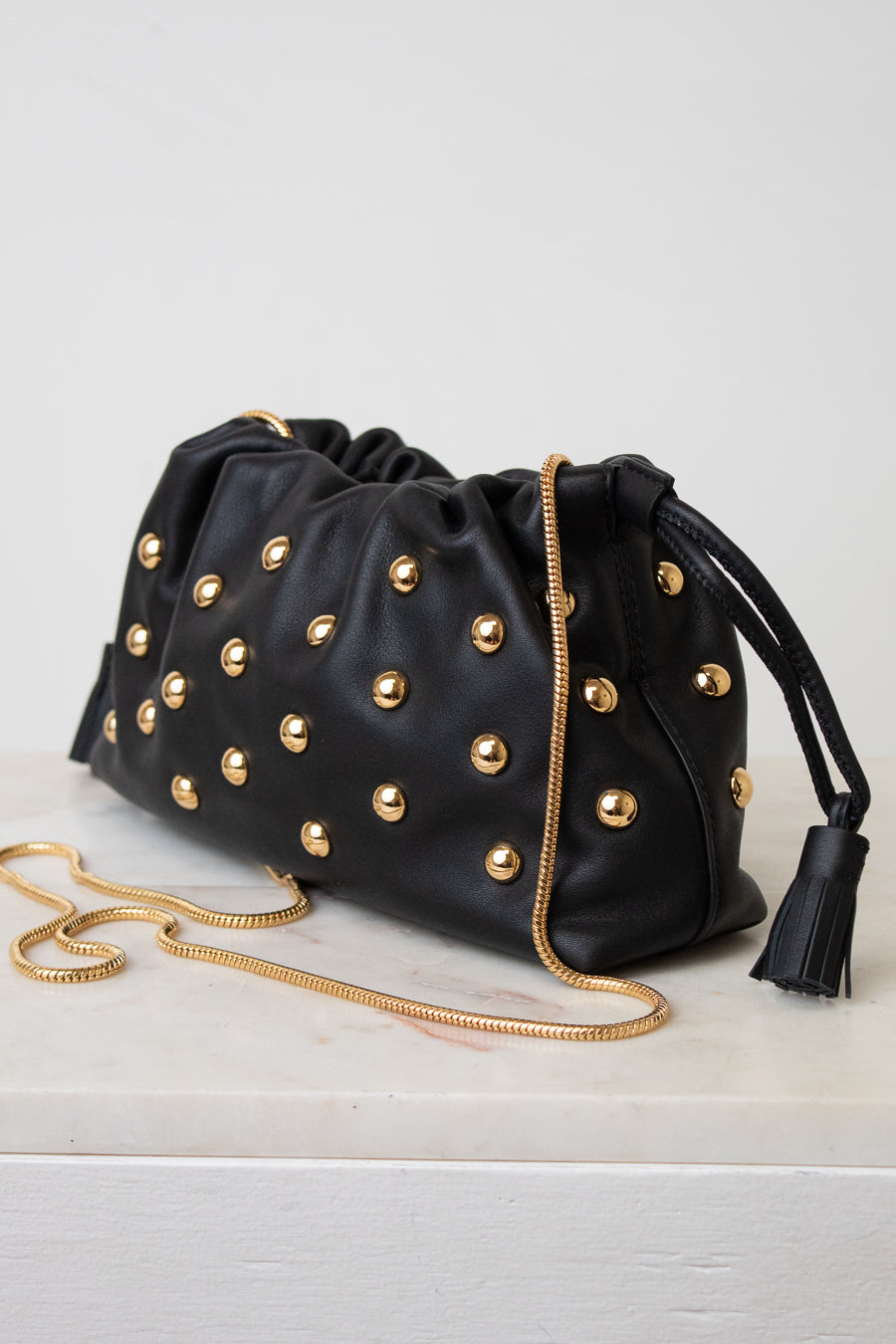 Black leather clutch bag with gold-tone studs, a gold chain strap, and a black tassel detail, displayed on a white surface against a light gray background.