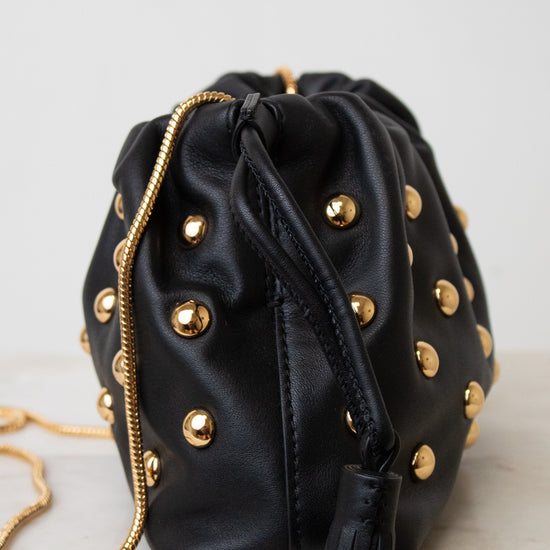 Black leather drawstring bucket bag with gold-tone studs and a gold chain strap, shown on a light neutral background.