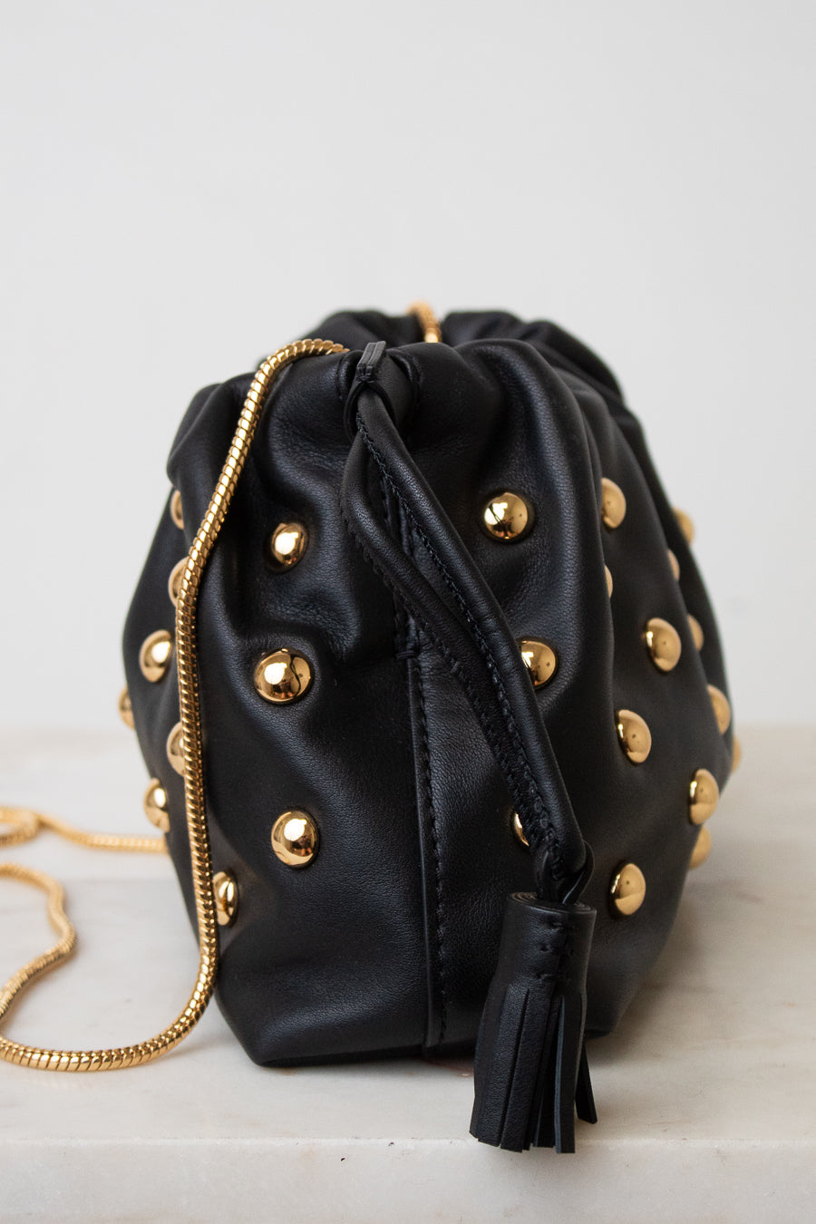 Black leather drawstring bucket bag with gold-tone studs and a gold chain strap, shown on a light neutral background.