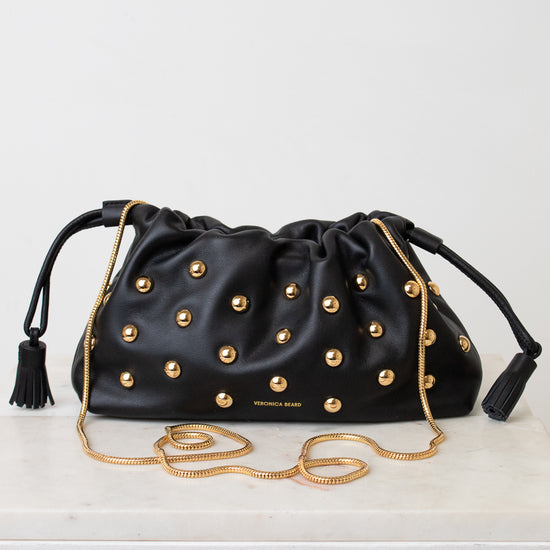 A black leather drawstring clutch bag by Veronica Beard, embellished with gold-tone studs and featuring a gold chain strap, resting on a white surface against a white background.