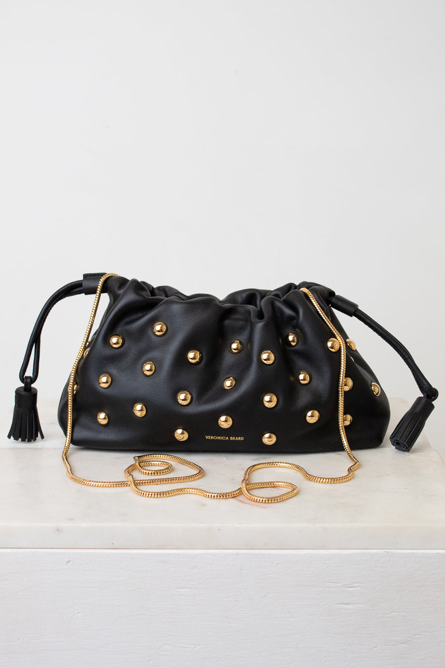 A black leather drawstring clutch bag by Veronica Beard, embellished with gold-tone studs and featuring a gold chain strap, resting on a white surface against a white background.