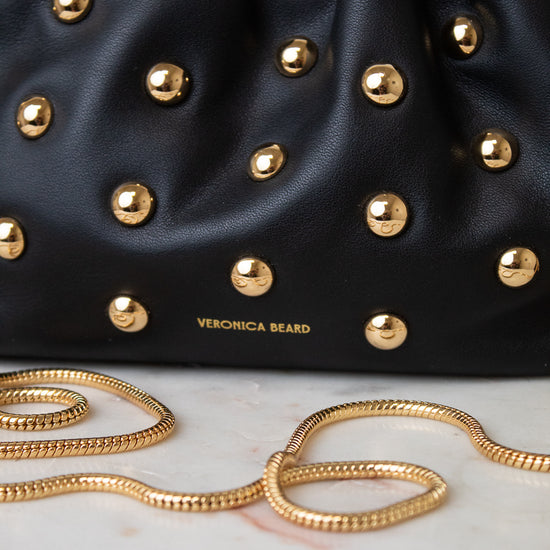 Close-up of a black leather handbag with gold studs and a gold chain strap, with the brand name 'VERONICA BEARD' printed in gold on the front.