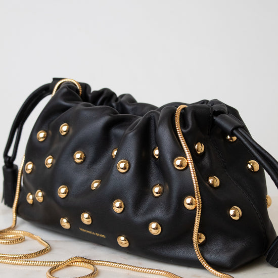 A black leather drawstring bag adorned with gold-tone studs and a gold chain strap, resting on a light marble surface against a neutral light gray background.