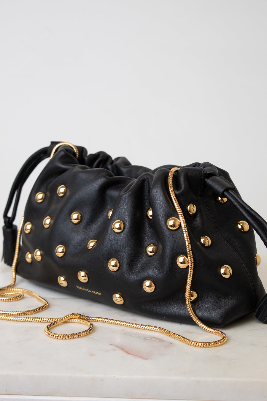 A black leather drawstring bag adorned with gold-tone studs and a gold chain strap, resting on a light marble surface against a neutral light gray background.